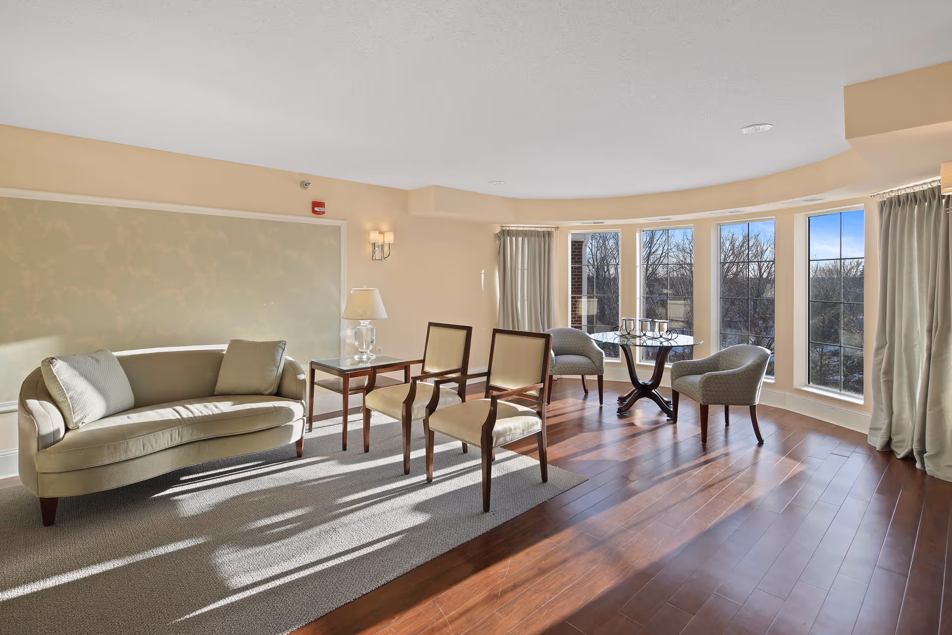 A bright and spacious living room area with a curved beige sofa, two wooden chairs with cream cushions, a glass table lamp on a side table, and a round glass table with two upholstered chairs near large windows letting in natural light.