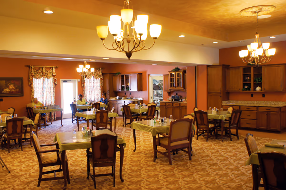 A dining room in a senior living facility with several tables covered with green tablecloths and wooden chairs. The room has warm orange walls, patterned carpet, and multiple chandeliers providing light. There are a few elderly people seated at the tables near the windows with floral curtains. Wooden cabinets and a countertop are visible along one wall.