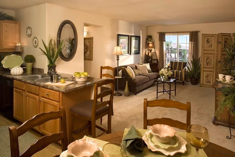 A cozy senior living apartment interior featuring a kitchen with wooden cabinets and a countertop with a sink, a dining table set with plates and glasses, and a living room area with a sofa, armchair, coffee table, floor lamp, and decorative plants. The room is warmly lit with natural light coming through a window with curtains.