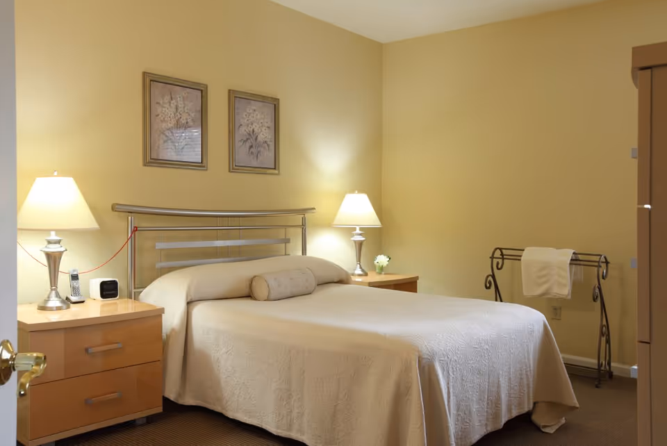 A neatly made bed with a light beige bedspread and a cylindrical pillow in a softly lit bedroom. There are two wooden nightstands on either side of the bed, each with a silver lamp. On the left nightstand, there is a cordless phone and a small clock. On the right nightstand, there is a small flower vase. Two framed floral pictures hang on the wall above the bed. A metal towel rack with a white towel is placed against the right wall. The walls are painted a warm yellow color.