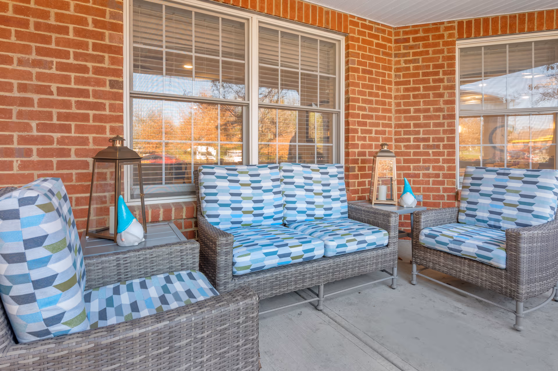 Outdoor seating area with three cushioned wicker chairs and a loveseat with blue, white, and green geometric patterned cushions. Two lanterns and two small decorative gnome figurines are placed on side tables. The area is adjacent to a brick wall with two large windows.