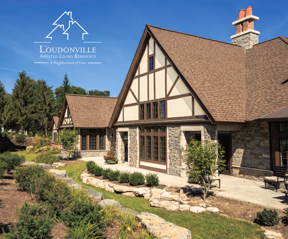 Exterior view of Loudonville Assisted Living Residence showing a building with stone and wood paneling, large windows, a brown shingled roof, and a landscaped garden with shrubs and trees under a clear blue sky.