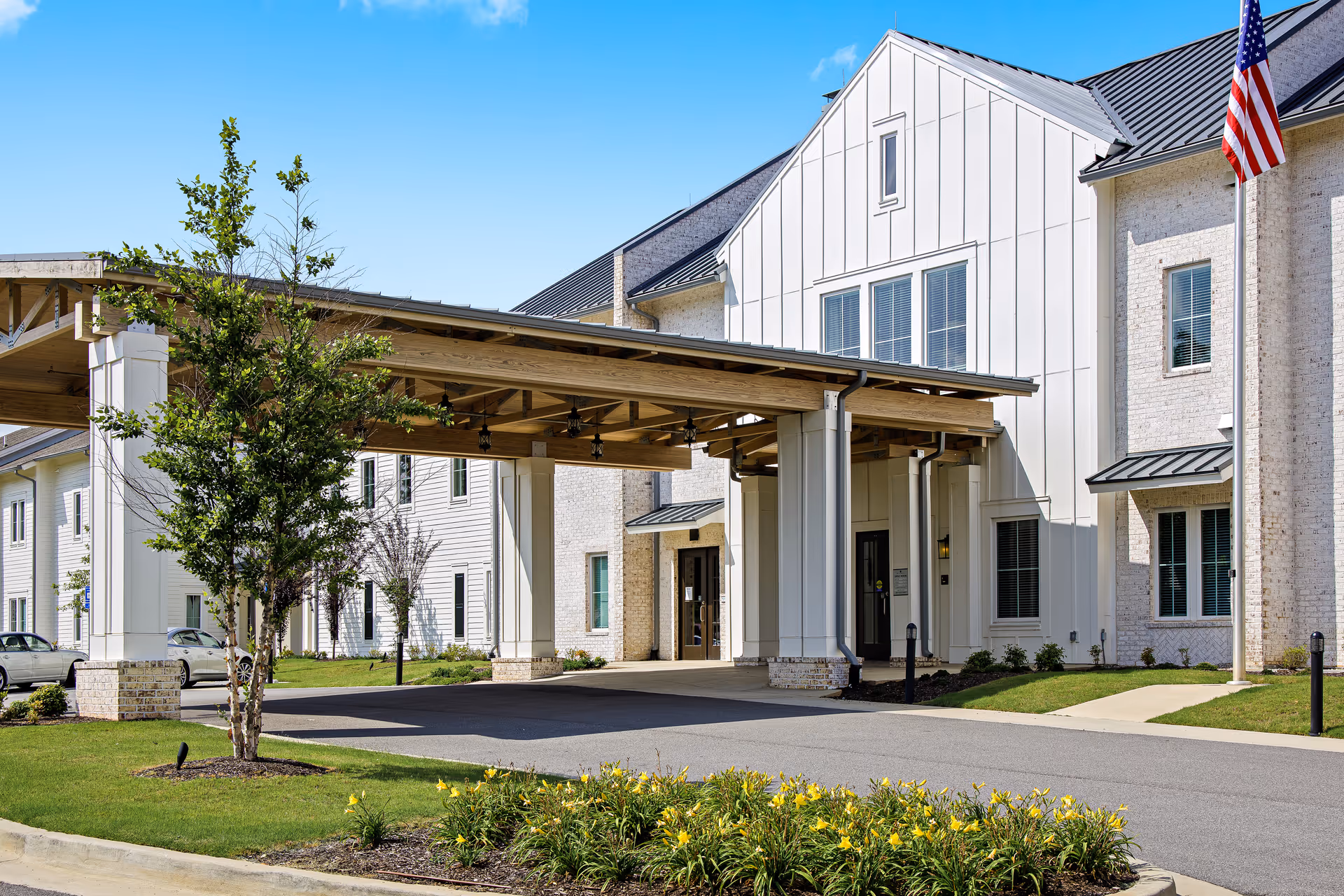Exterior view of The Claiborne at Gulfport Highlands facility showing a covered entrance with wooden beams, white brick and siding walls, windows, a small tree, and a flower bed with yellow flowers under a clear blue sky.