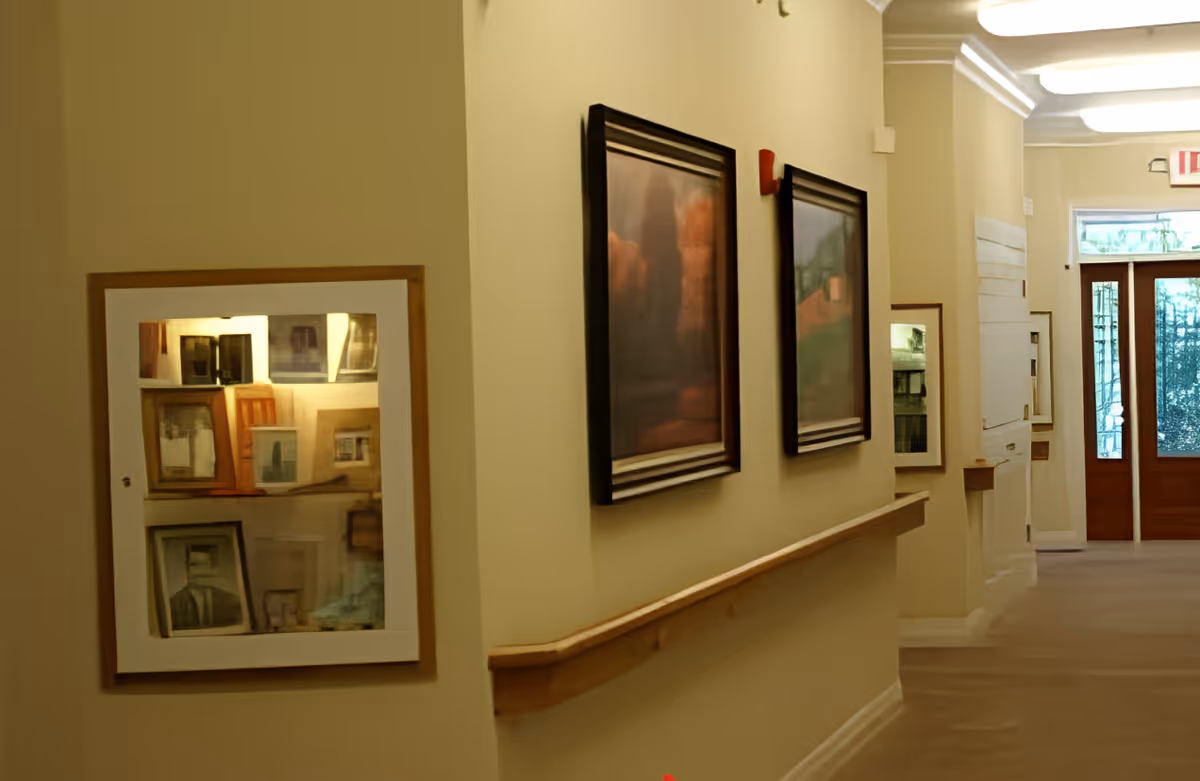 A well-lit hallway in a senior living facility with beige walls and carpeted floor. The hallway features a wooden handrail along the right wall and several framed pictures hanging above it. At the end of the hallway, there is a wooden door with glass panels allowing natural light to enter. On the left wall, there is a display case with various framed photos or artwork inside.