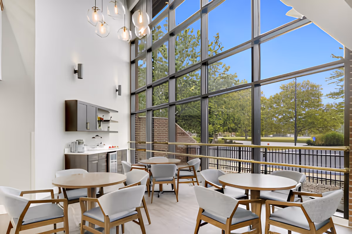 A bright dining area with round wooden tables and cushioned chairs. The space features a large floor-to-ceiling window wall offering a clear view of green trees and a blue sky outside. There is a small kitchenette with cabinets, a countertop, and a wine cooler along one wall. Modern pendant lights hang from the ceiling.