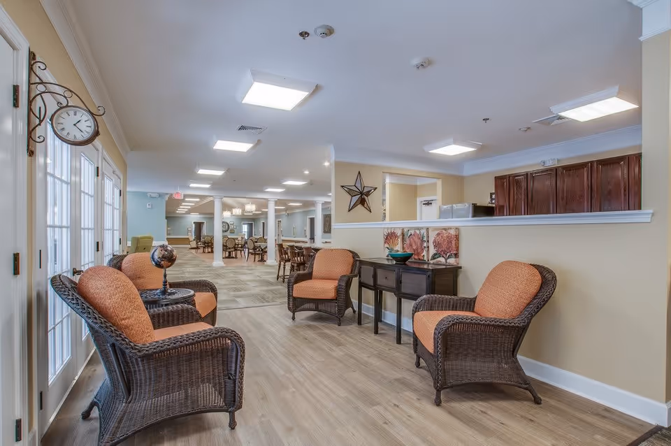 A bright and spacious common area in Barton House Memory Care featuring wicker chairs with orange cushions arranged around a small table with a decorative globe. The room has light wood flooring, beige walls, and ceiling lights. In the background, there are dining tables and chairs, white columns, and a decorative star on the wall.