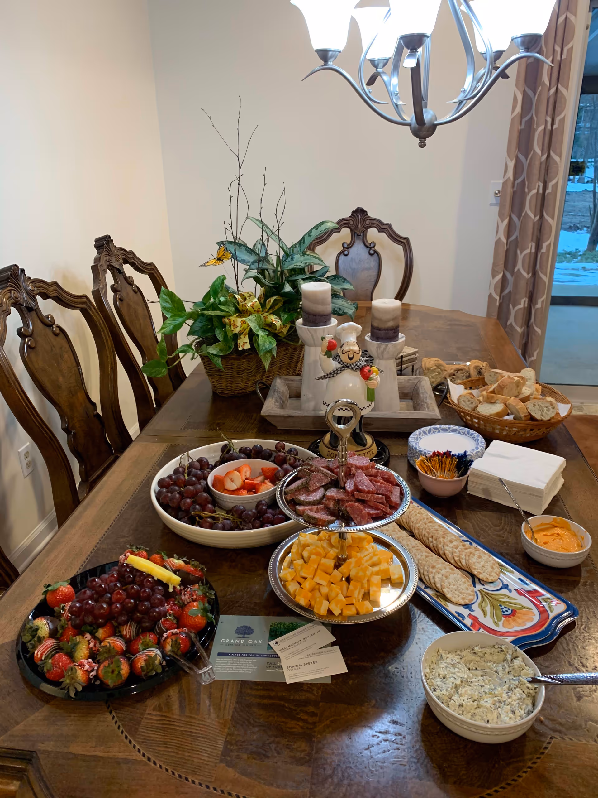 A dining table set with a variety of snacks including grapes, strawberries, cheese cubes, sliced salami, crackers, bread, and dips. There is a decorative plant and a chef figurine holding vegetables in the center of the table. Wooden chairs surround the table, and a chandelier hangs above. A sliding glass door with curtains is visible in the background.
