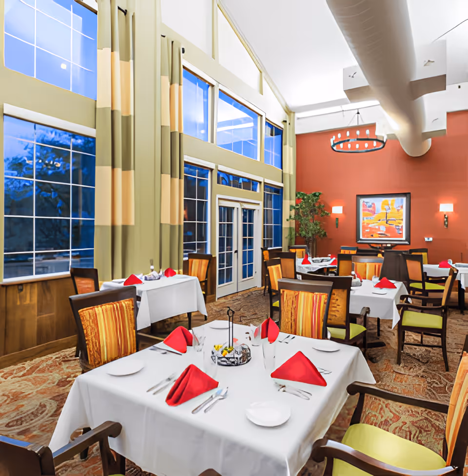 A bright and elegant dining room with large windows and high ceilings. Tables are set with white tablecloths, red folded napkins, plates, and silverware. The room features patterned chairs, a red accent wall with a colorful painting, wall sconces, and a large circular chandelier. There is a potted plant in the corner and a carpeted floor with intricate designs.