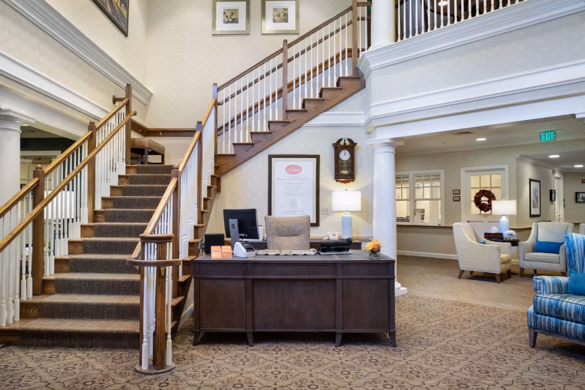 Lobby with a front desk in front of a carpeted staircase and a seating area beneath an upper balcony.