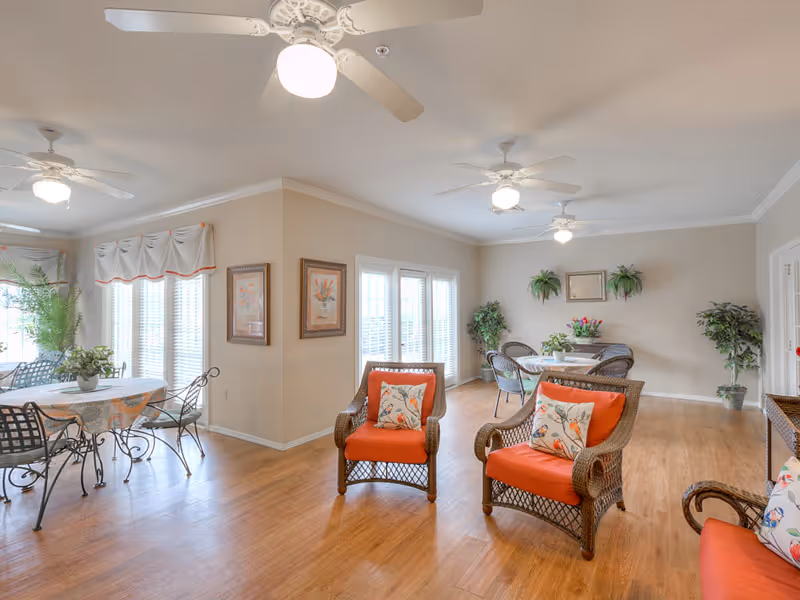 A bright and spacious living room area with wooden flooring, wicker chairs with orange cushions and floral pillows, round tables with chairs, ceiling fans with lights, large windows with white blinds, and potted plants and framed artwork on the walls.