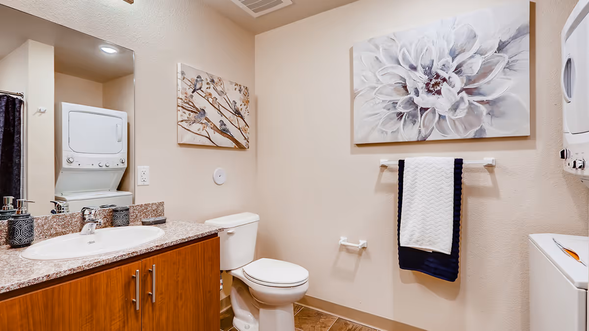 A bathroom with a white toilet, a wooden vanity with a granite countertop and a sink, a large mirror above the sink, and a stacked washer and dryer unit. The walls are decorated with two paintings, one of birds on a branch and another of a large white flower. A towel rack holds a white and navy blue towel.