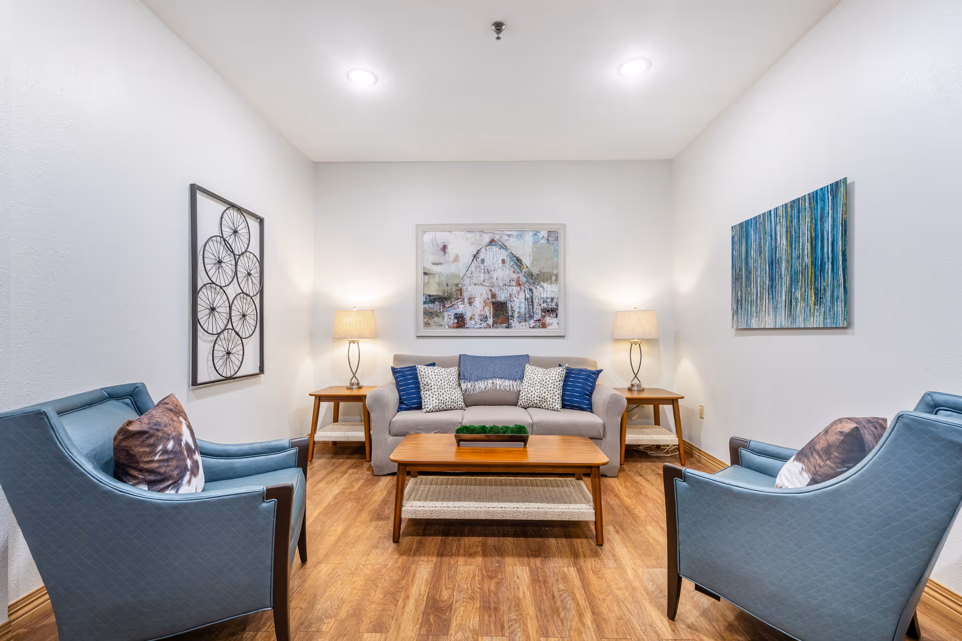 A cozy living room with a beige sofa adorned with blue and patterned pillows, two blue armchairs with brown and white pillows, a wooden coffee table with a green centerpiece, two wooden side tables each with a lamp, and three pieces of wall art including a painting of a barn and abstract designs.
