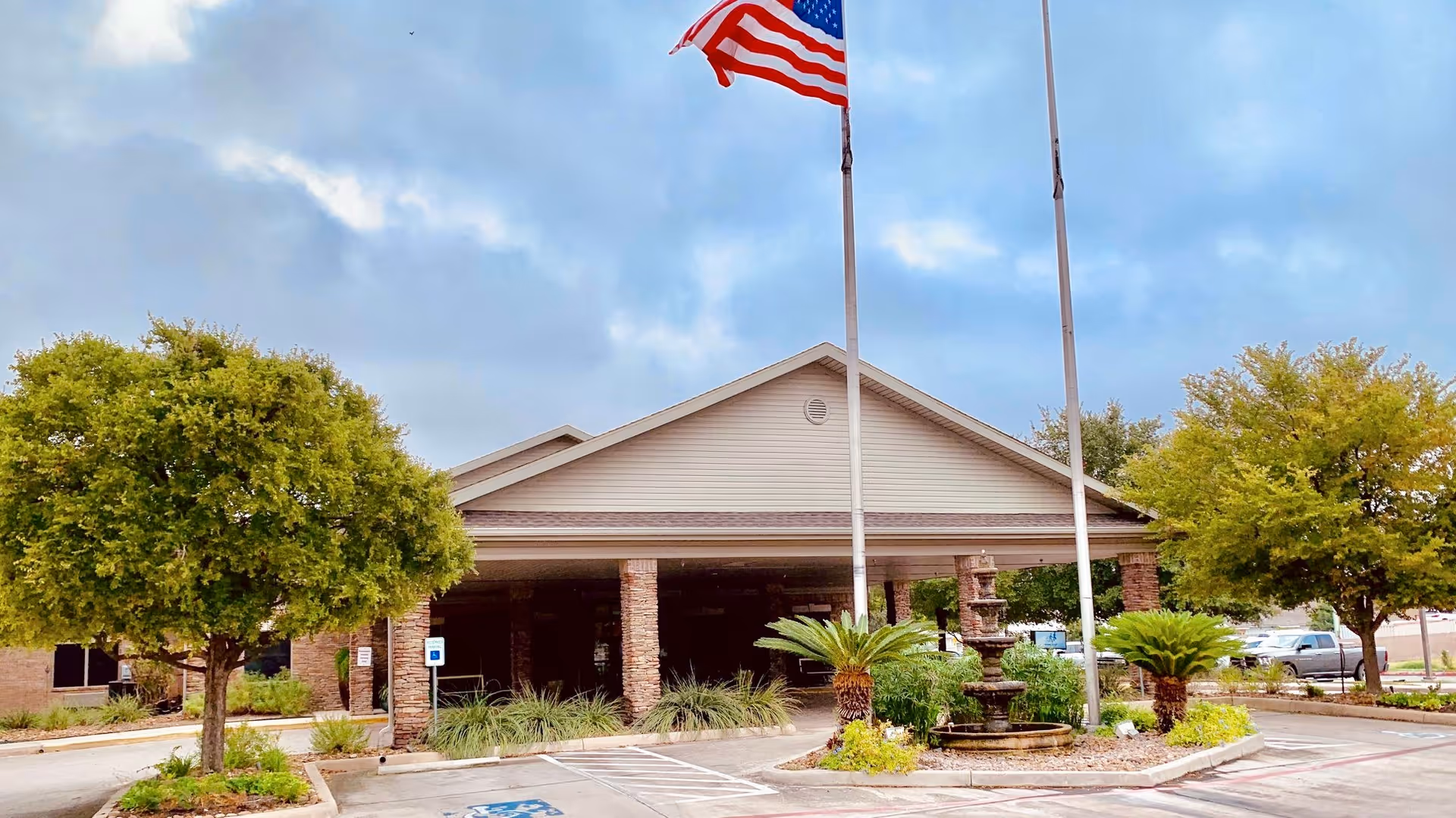 Front exterior view of Silver Tree Nursing and Rehabilitation facility with an American flag on a flagpole, two trees, a fountain, and a parking area with handicap parking spaces.