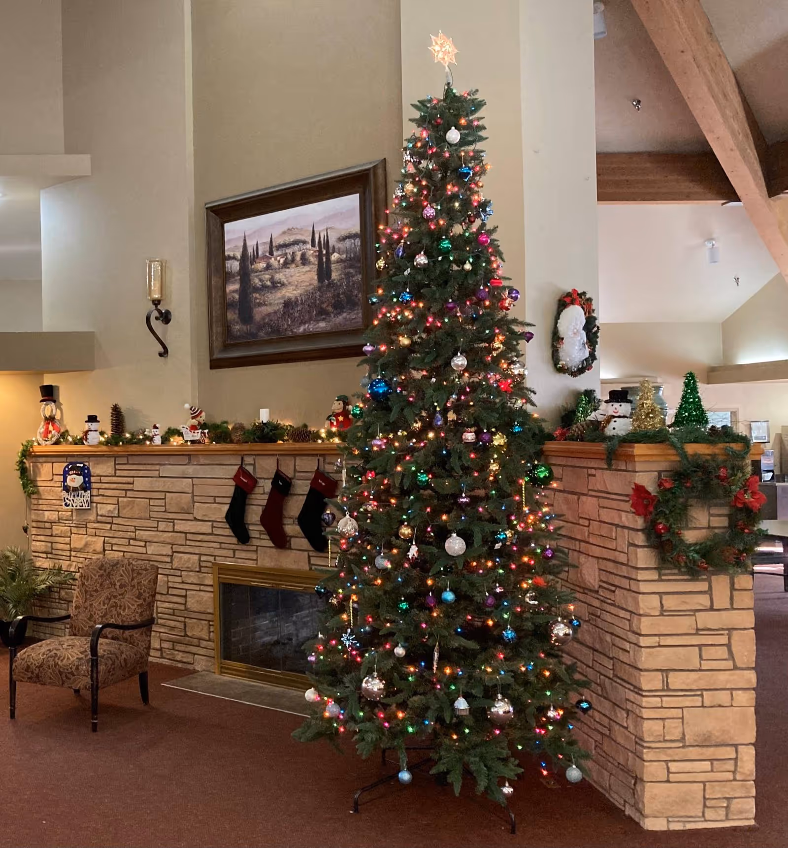 A tall, decorated Christmas tree with colorful lights and ornaments stands in a cozy room next to a stone fireplace adorned with holiday stockings, garlands, and festive decorations. A patterned armchair is placed near the fireplace, and a framed landscape painting hangs on the wall above it. Additional holiday wreaths and decorations are visible on the walls and mantel.