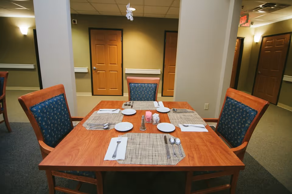 A wooden dining table set for four with placemats, plates, napkins, and cutlery in a room with beige walls and multiple closed wooden doors. The chairs have wooden frames with blue patterned cushions.