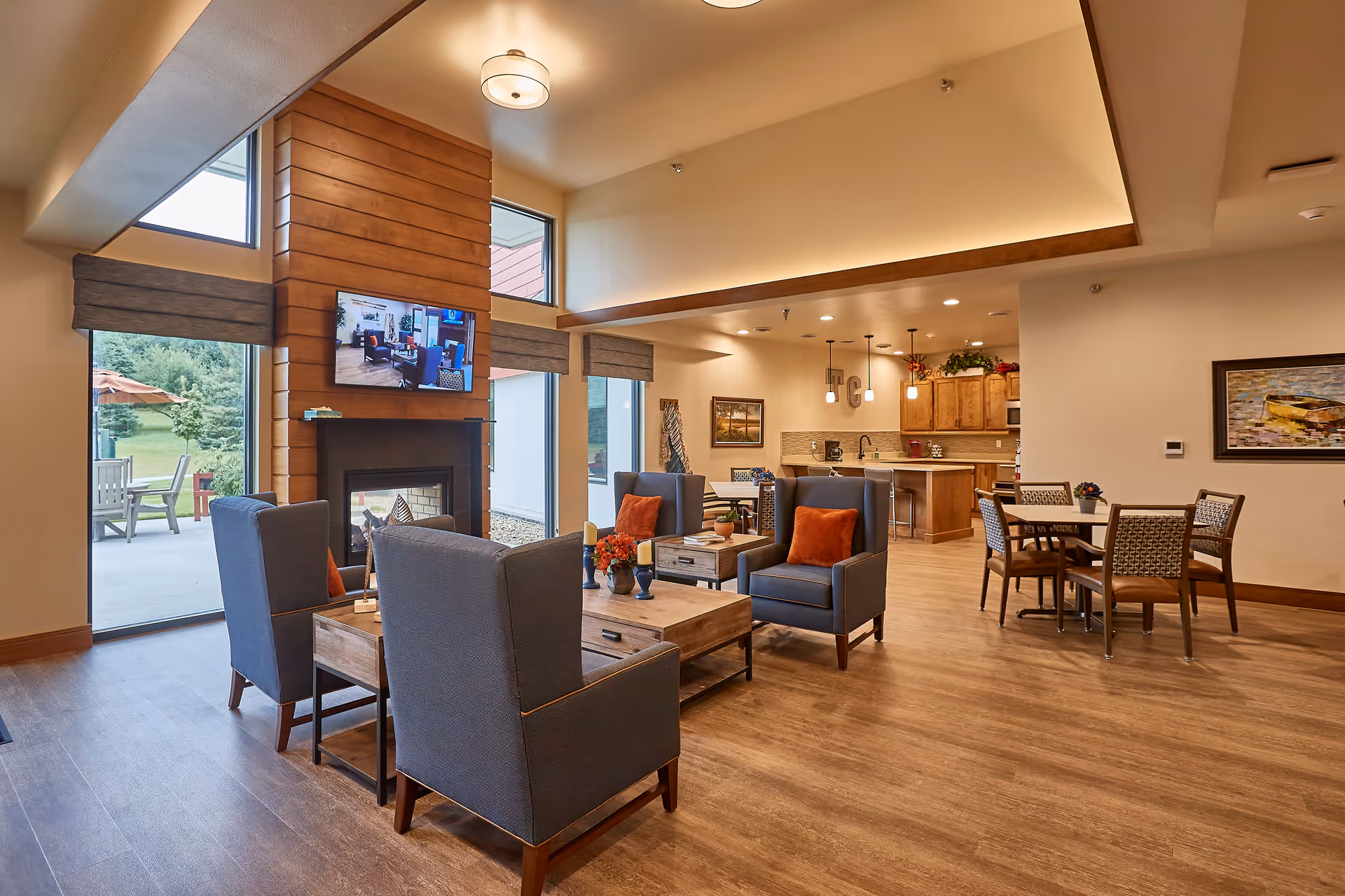 A spacious and well-lit senior living common area featuring four blue armchairs with orange pillows arranged around a wooden coffee table in front of a modern fireplace with a mounted flat-screen TV above it. Large windows and glass doors provide a view of an outdoor patio with chairs and umbrellas. In the background, there is a dining area with tables and chairs and a kitchen with wooden cabinets and pendant lights.