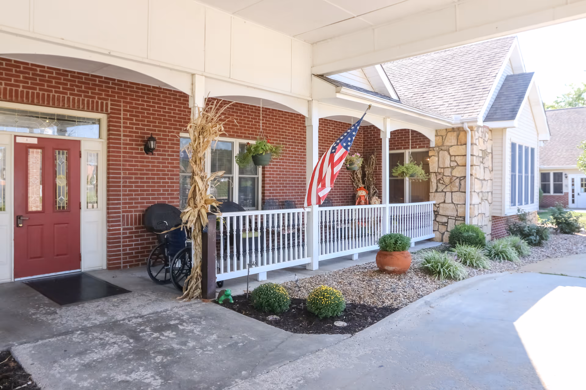 Covered entrance area of a senior living facility with a red door, brick walls, white railing, hanging plants, an American flag, and a wheelchair near the door. There are decorative plants and landscaping along the walkway.