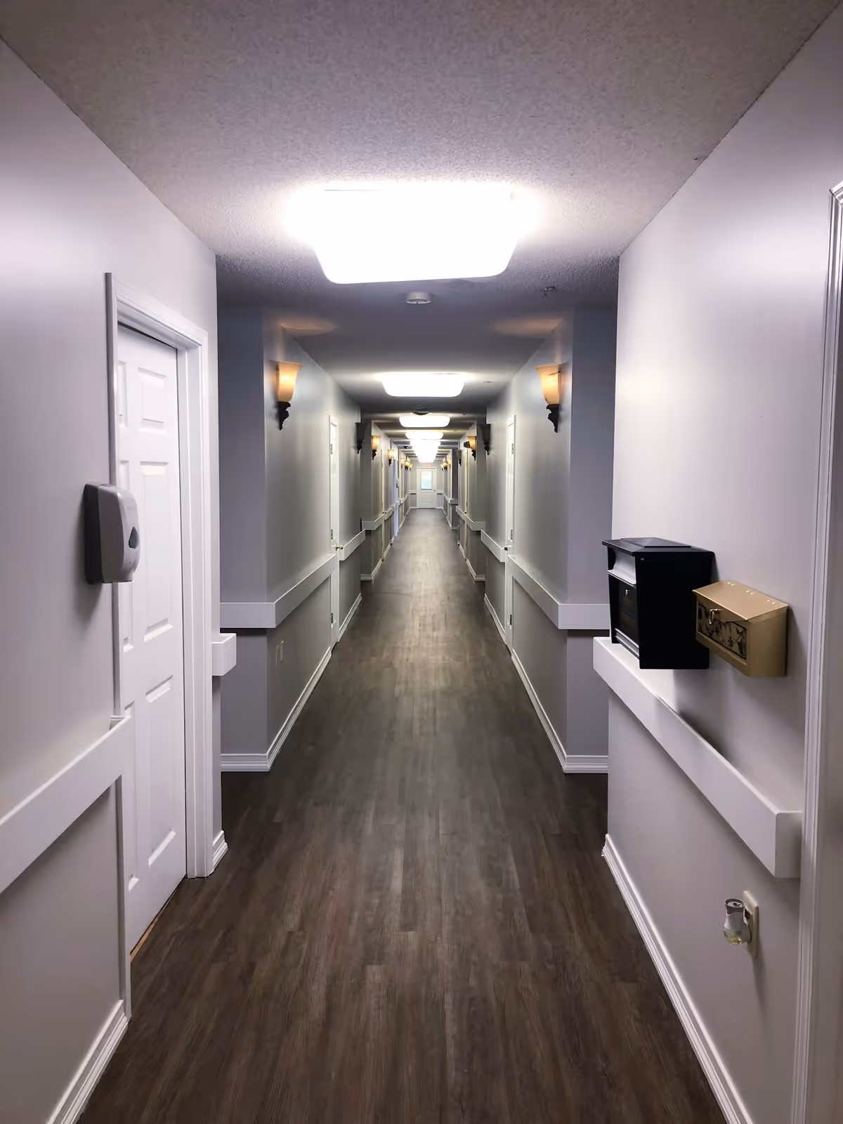 A long, well-lit hallway in a senior living facility with white walls, wooden flooring, and multiple doors on both sides. The hallway has wall-mounted lights and ceiling lights evenly spaced along the length.
