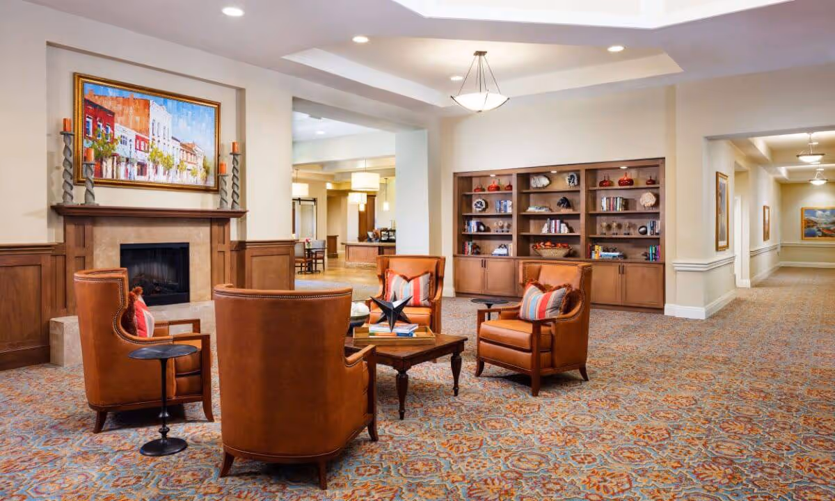 A cozy seating area in a senior living facility with four brown leather armchairs arranged around a wooden coffee table. The room features a fireplace with a colorful painting above it, built-in wooden bookshelves filled with decorative items and books, and a patterned carpet. The space is well-lit with ceiling lights and has a hallway leading to other parts of the facility.