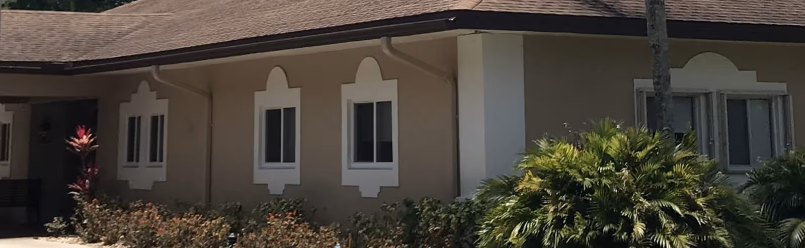 Exterior view of a single-story building with beige walls and a brown shingled roof. The building has several white-framed windows with decorative trim. There are bushes and plants, including a tall red-leafed plant and green palm-like shrubs, in front of the building.