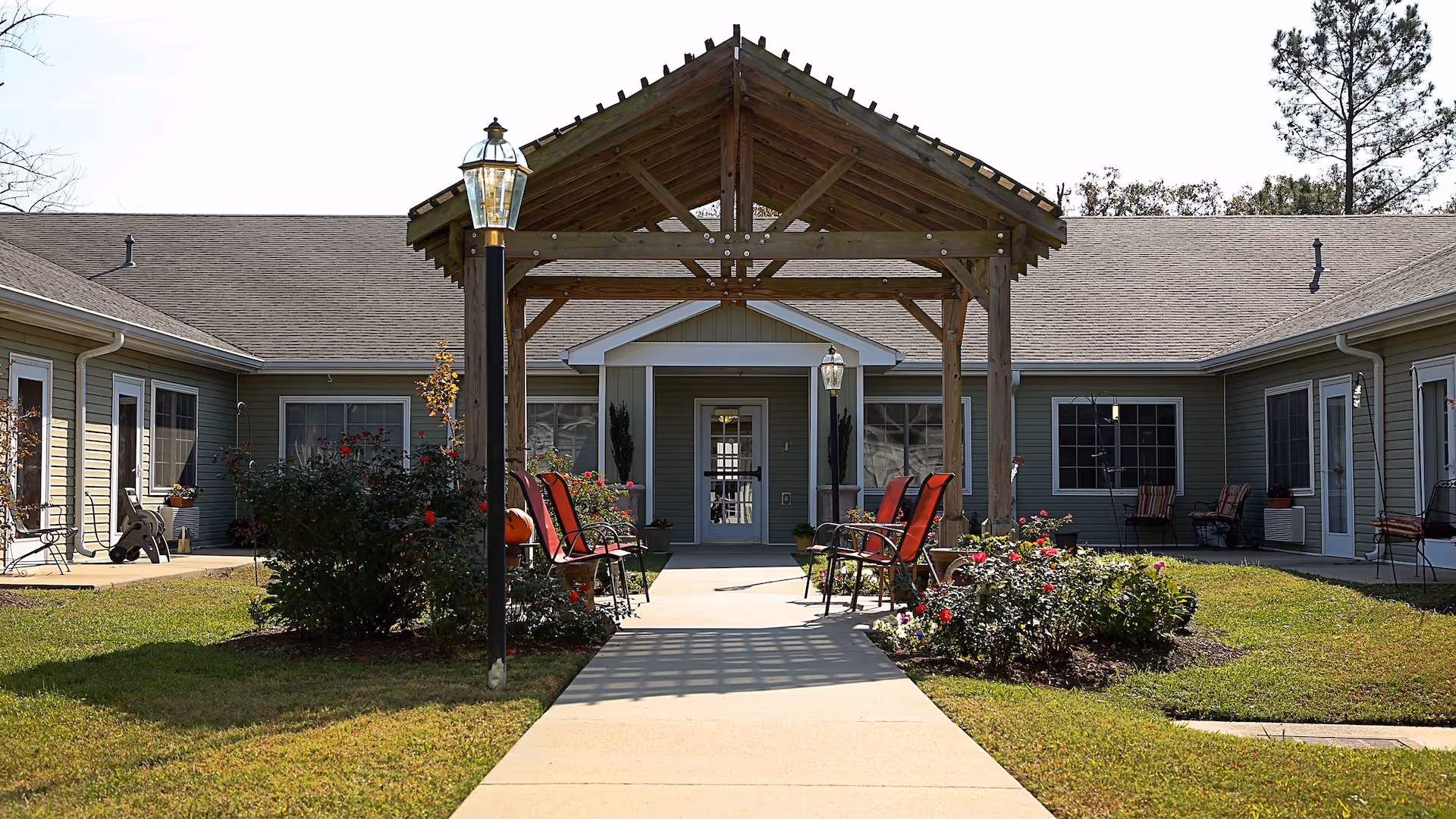 Outdoor courtyard area of a senior living facility with a wooden pergola, red chairs arranged under it, flower beds on either side of the walkway, and a single-story building with green siding surrounding the courtyard.