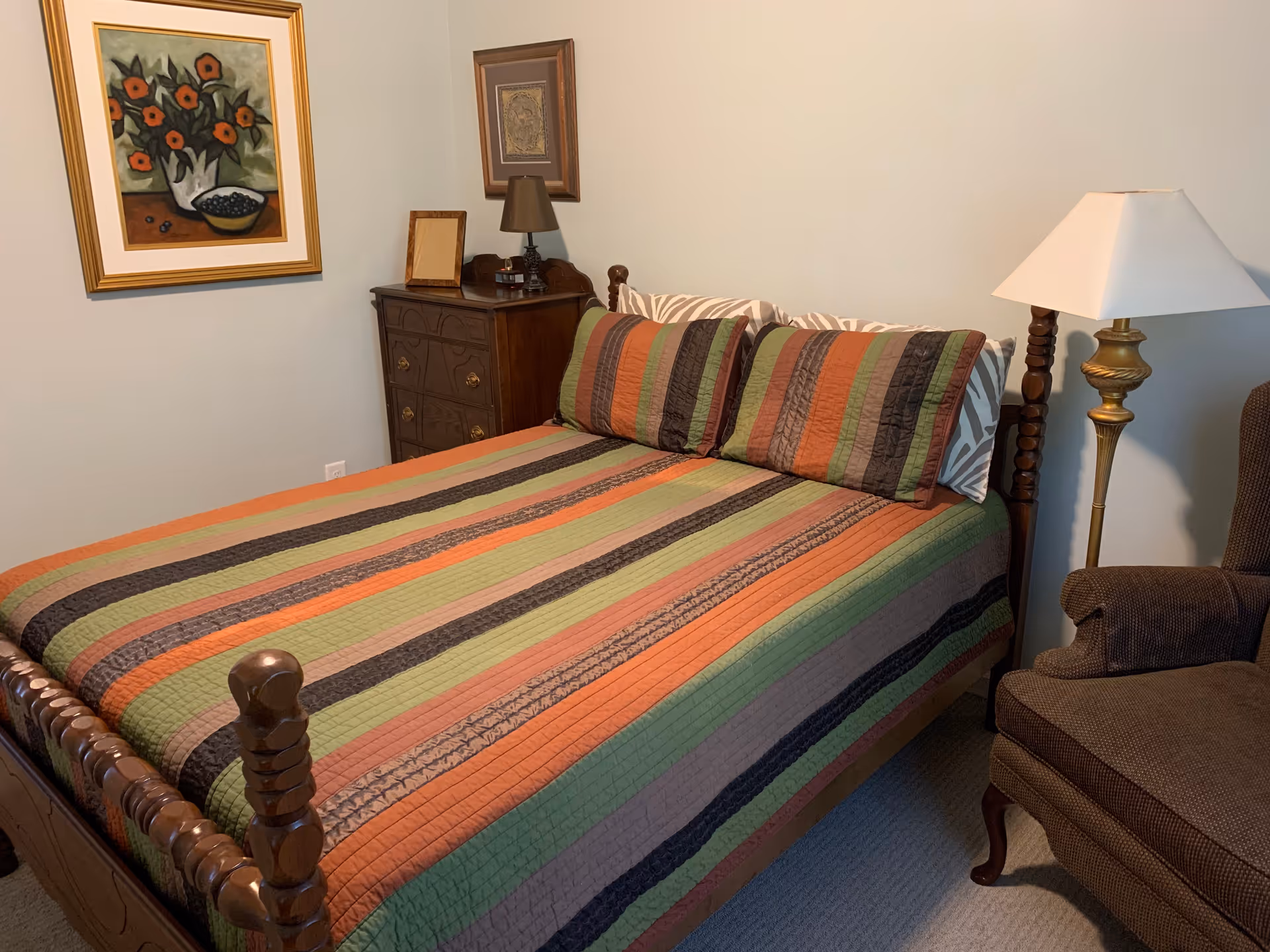 Neatly made bedroom with a striped multicolored bedspread on a wooden bedframe, a nightstand with a lamp and framed art on the wall, and an upholstered chair.