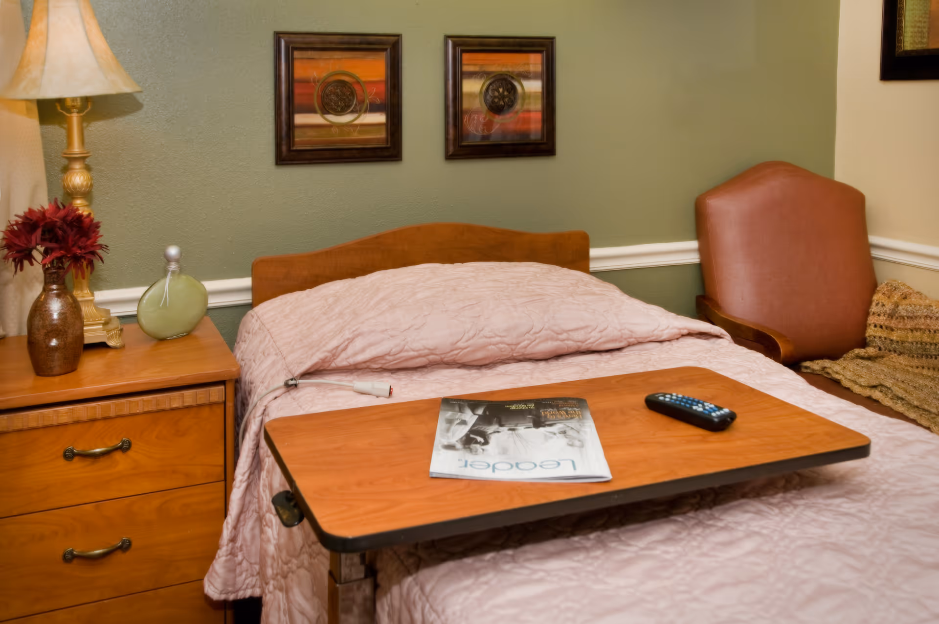 A bedroom in a senior living facility with a single bed covered in a light pink quilt. A wooden overbed table holds a magazine and a TV remote. Next to the bed is a wooden nightstand with a lamp, a vase with red flowers, and a decorative item. A brown upholstered chair and a knitted blanket are visible in the corner. Two framed artworks hang on the green wall behind the bed.