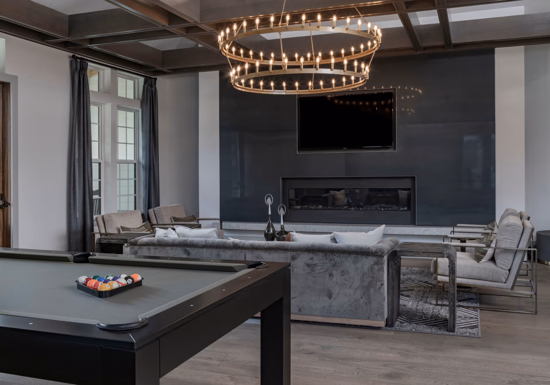 Modern common lounge with a pool table in the foreground, gray sofas and armchairs facing a wall-mounted TV and fireplace beneath a large circular chandelier.