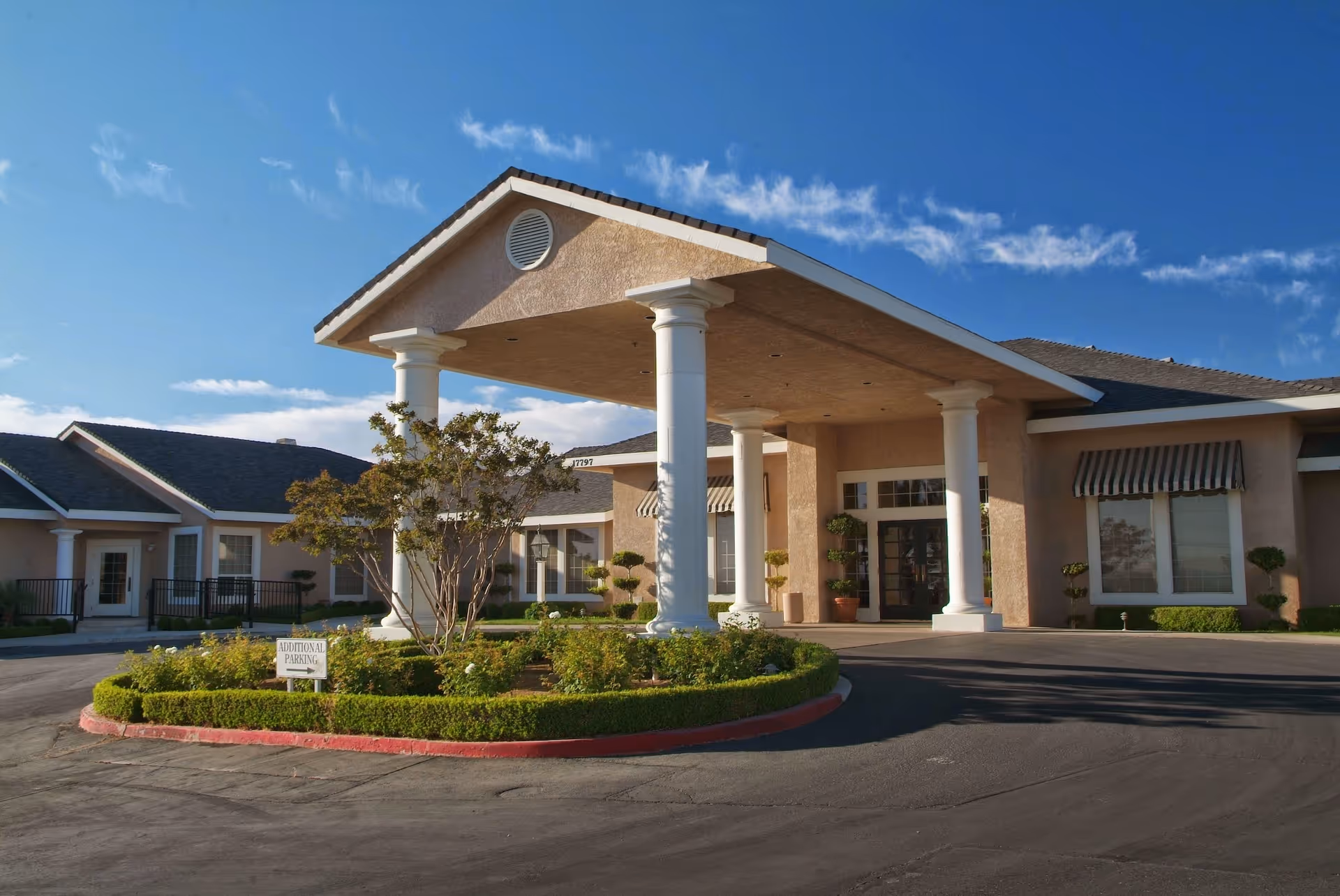Exterior view of Sterling Commons Memory Care Community building with a covered entrance supported by white columns, landscaped greenery, and a clear blue sky.
