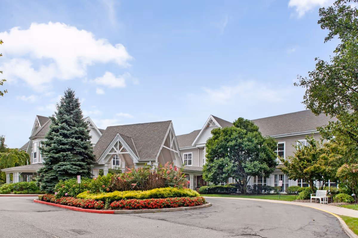 Exterior view of a senior living facility named Sunrise of Holbrook, featuring a large building with peaked roofs, surrounded by well-maintained landscaping including trees, bushes, and colorful flower beds under a partly cloudy sky.