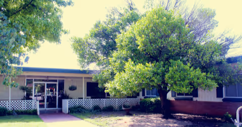 Front entrance of a single-story care facility with a walkway, white lattice railing, and a large tree on the lawn.