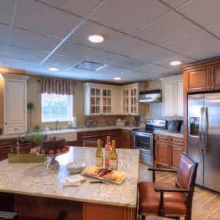 A modern kitchen with a large granite island countertop in the center, featuring two bottles of wine, a plate of cheese and crackers, and a bowl of fruit. The kitchen has wooden cabinets in two tones, stainless steel appliances including a refrigerator and oven, and a window with a valance letting in natural light.