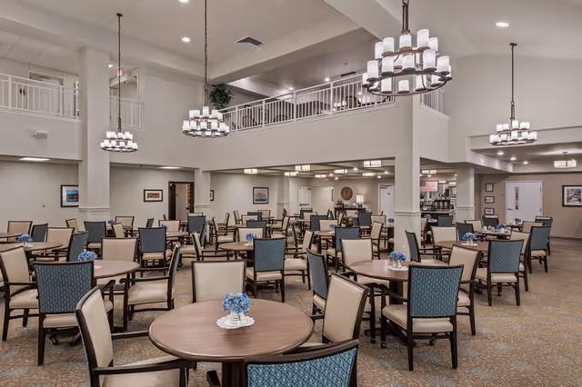 A spacious dining room with multiple round wooden tables and chairs arranged neatly. Each table has a small floral centerpiece with blue flowers. The room features high ceilings with modern chandeliers hanging down, beige walls, and a carpeted floor. There is a second-floor balcony overlooking the dining area.