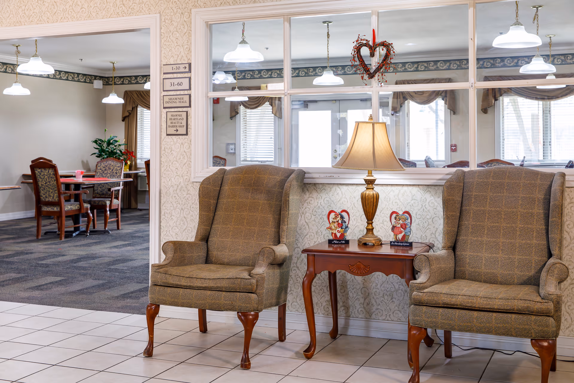 Two upholstered armchairs flanking a wooden side table with a lamp in a senior living facility common area, with a dining area visible through a glass partition.