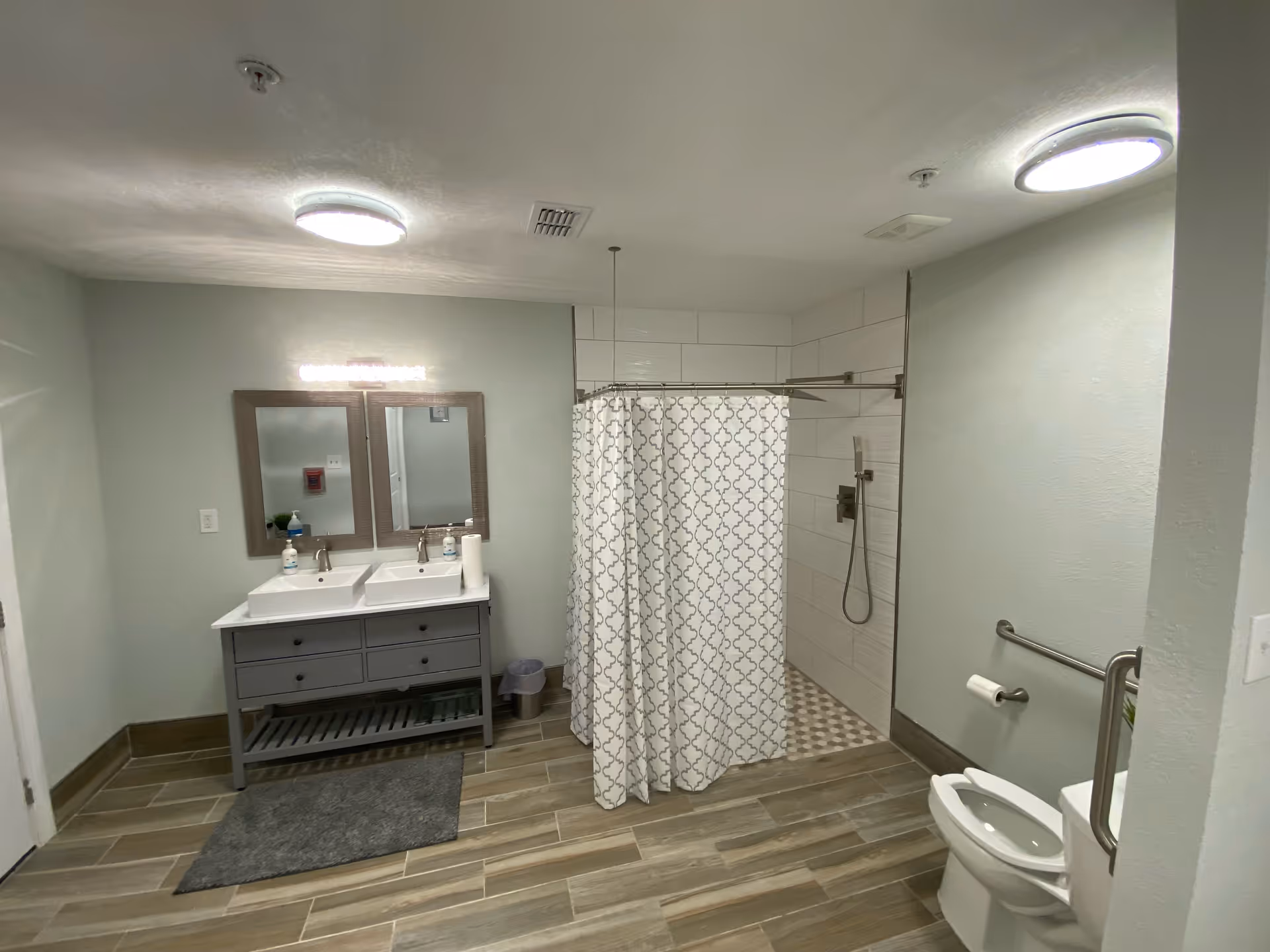 A spacious bathroom with two sinks and mirrors on the left, a shower area with a patterned curtain in the center, and a toilet with grab bars on the right. The floor has wood-like tiles and the walls are painted light green. The room is well-lit with ceiling lights.