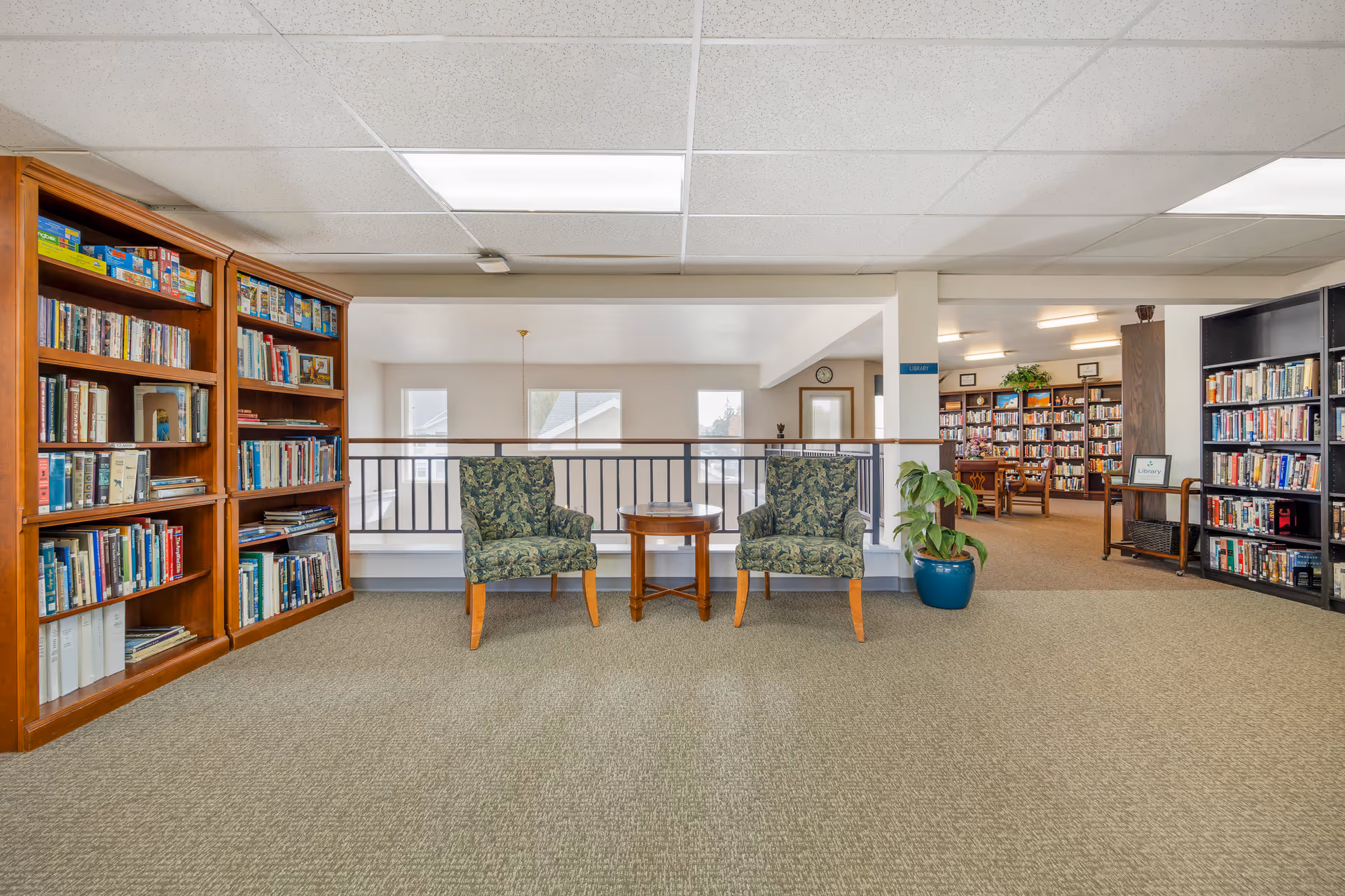 Interior view of a library or reading area in a senior living facility with bookshelves filled with books on both sides. Two patterned armchairs with wooden legs are positioned around a small round wooden table. A potted plant is placed next to one of the chairs. The area is carpeted and well-lit with ceiling lights.