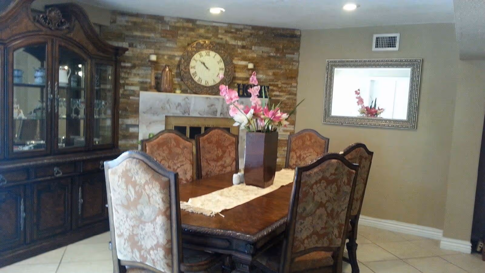 A dining room with a wooden dining table and six upholstered chairs. A vase with pink and white flowers is placed on a table runner in the center of the table. Behind the table is a stone fireplace with a large round clock above it. To the left is a wooden china cabinet with glass doors displaying dishes and glassware. A large framed mirror hangs on the beige wall to the right.