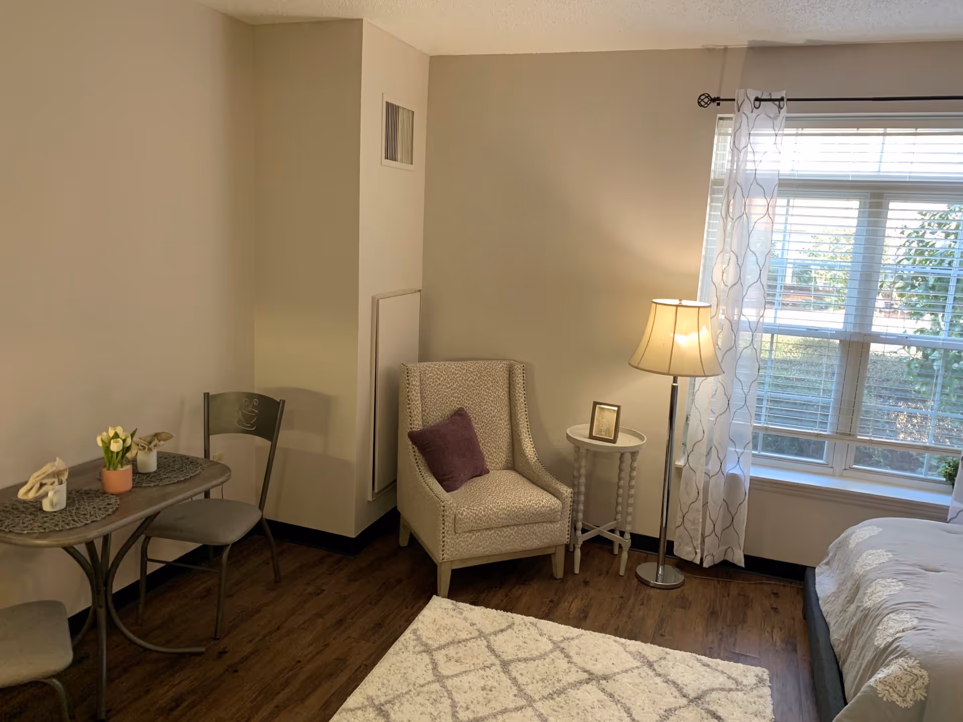 A cozy senior living room with a small dining table and two chairs on the left, a patterned armchair with a purple pillow next to a small round side table with a framed photo, a floor lamp, and a window with white curtains letting in natural light. The floor is wooden with a white patterned rug partially visible.