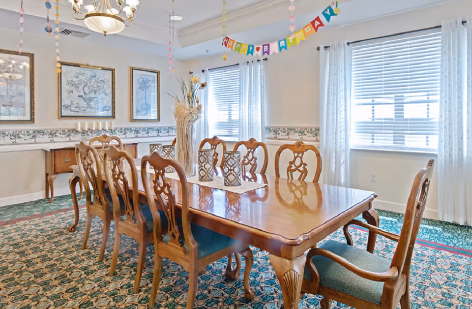 A bright dining room with a long wooden table surrounded by eight wooden chairs with green cushions. The room has patterned carpet, three framed pictures on the wall, two windows with white curtains, and a 'Happy Birthday' banner hanging from the ceiling. Decorative vases and candles are placed on the table and sideboard.
