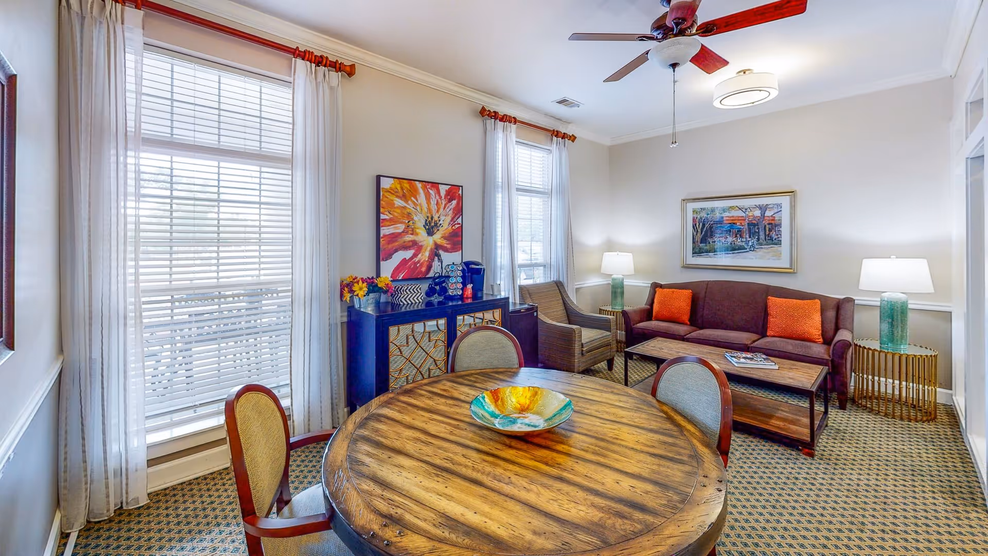 A cozy living room area with a round wooden table and four chairs in the foreground. Behind the table is a dark brown sofa with two orange cushions, a wicker armchair, and two green glass table lamps on either side of the sofa. The room has two large windows with white curtains, a ceiling fan with wooden blades, and colorful artwork on the walls.