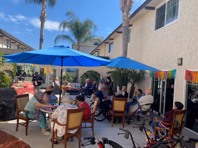A group of elderly people sitting and socializing outdoors under blue umbrellas in a courtyard area with palm trees and a beige building in the background. Some individuals are using wheelchairs and walkers. Colorful decorations hang on the building wall.
