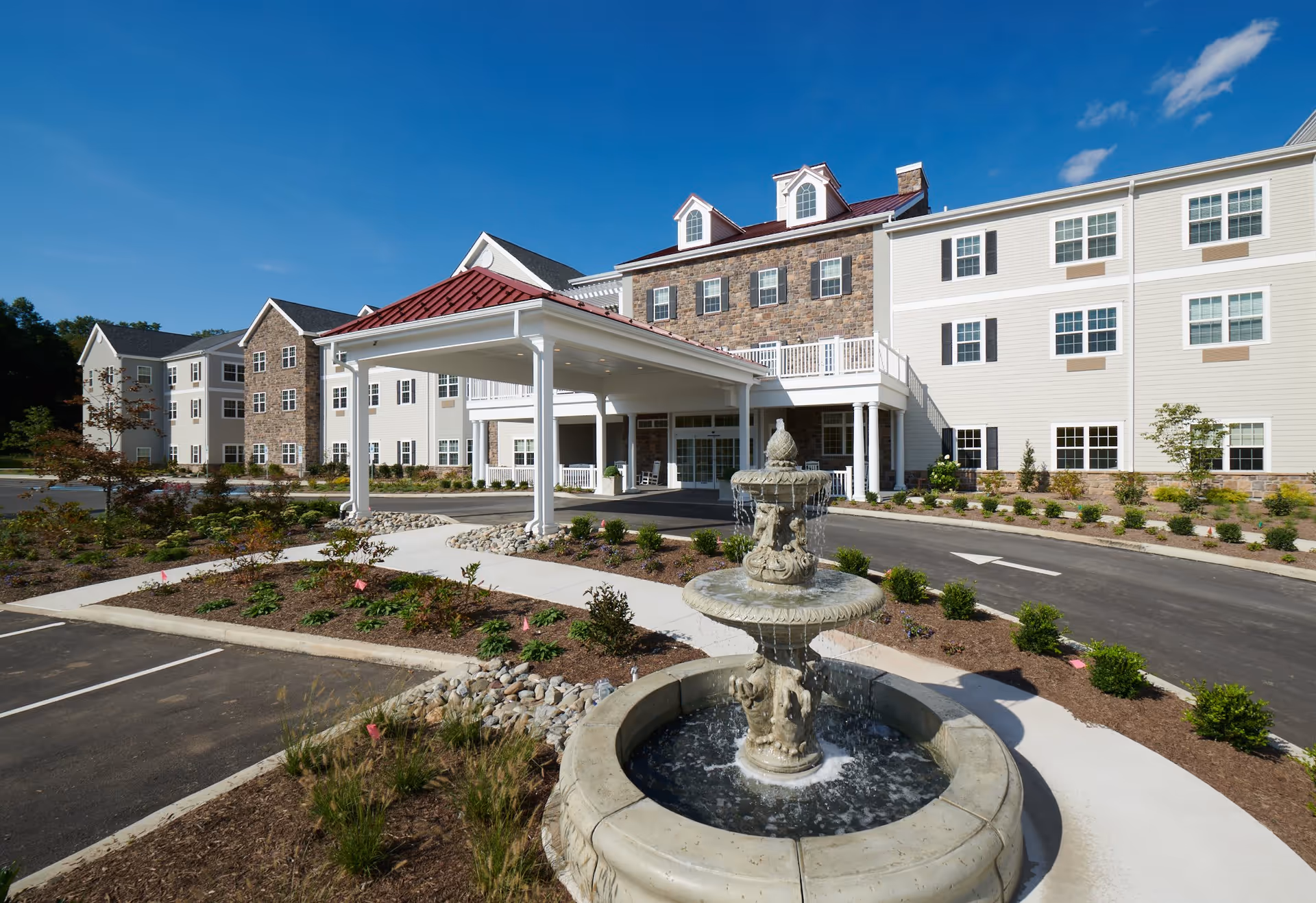 Exterior view of a senior living facility named The Village at Lifequest, featuring a large multi-story building with stone and siding facade, a covered entrance with white columns, landscaped garden beds, a circular driveway, and a decorative water fountain in the foreground under a clear blue sky.