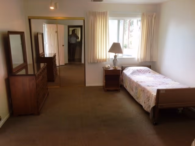 Bedroom in Crestview Manor with single bed, dresser, nightstand, lamp, and window.