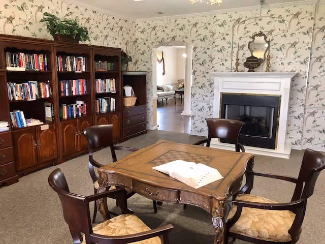 A cozy living room with a wooden square table surrounded by four cushioned wooden chairs. Behind the table is a white fireplace with a decorative mirror and an urn on the mantel. To the left, there is a large wooden bookshelf filled with books and a plant on top. The walls are decorated with floral wallpaper, and an open doorway leads to another room with additional seating.