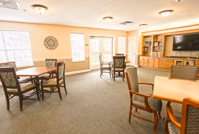 A well-lit common room with several wooden tables and cushioned chairs arranged around the room. The walls are painted a light beige color, and there are two windows and a set of French doors letting in natural light. On one side, there is a built-in wooden cabinet with shelves holding books and a large flat-screen TV mounted above it. The room has a carpeted floor and ceiling lights.