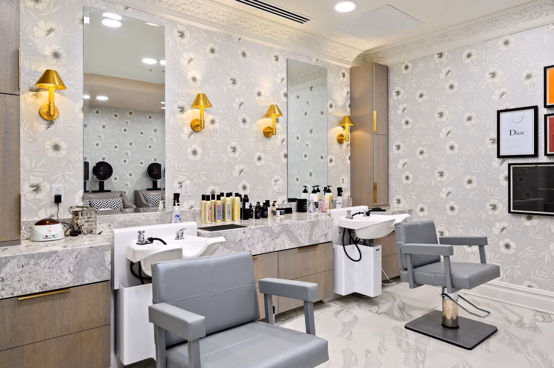 A modern salon area with two gray salon chairs in front of two white hair washing sinks. The wall behind features floral wallpaper, two large mirrors, and gold wall sconces. Various hair care products are arranged on a marble countertop beneath the mirrors. Framed artwork is visible on the right side of the wall.