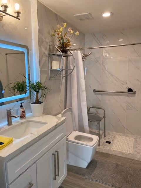 Well-lit accessible bathroom with a vanity and illuminated mirror, toilet, and a walk-in shower with grab bars and a shower chair.