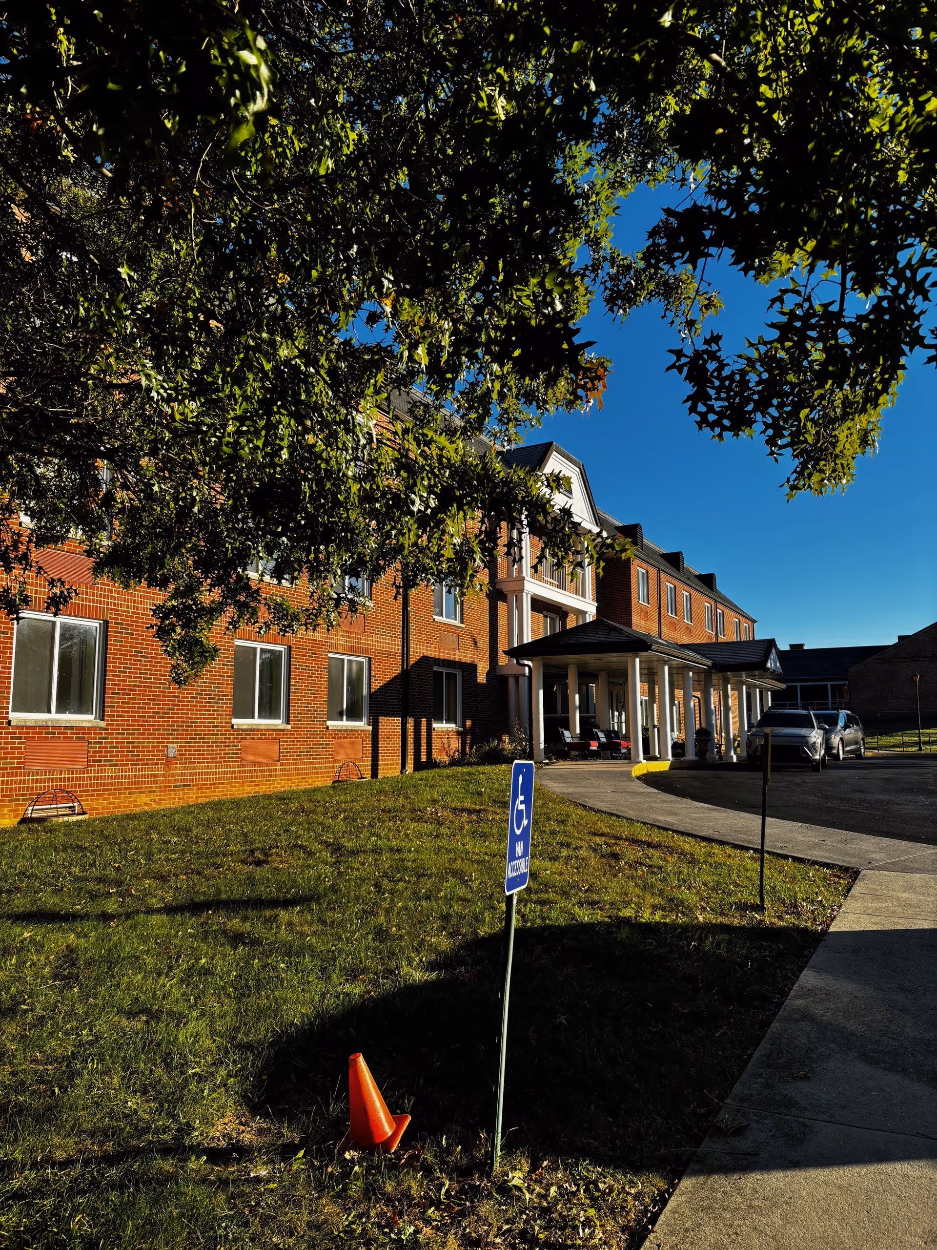 Exterior view of a brick senior living facility building with a covered entrance and a driveway. There is a handicapped parking sign on a grassy area in front of the building, with a small orange traffic cone nearby. Trees with green leaves partially frame the top of the image under a clear blue sky.