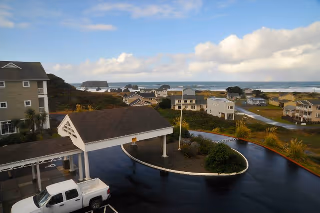 View of a senior living community with a covered driveway entrance, a white pickup truck parked nearby, multiple residential buildings, and a scenic ocean with rock formations in the background under a partly cloudy sky.