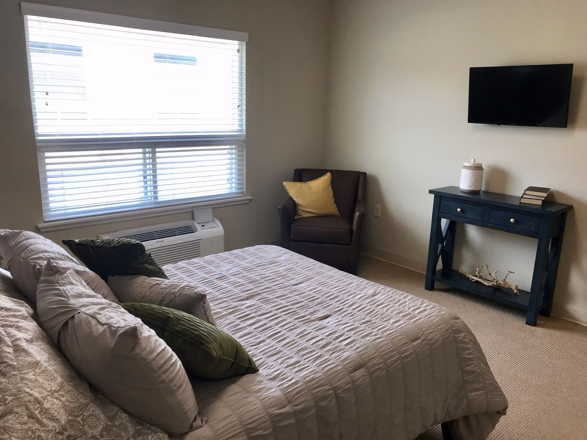 A bedroom with a neatly made bed featuring multiple pillows, a window with blinds, a brown armchair with a yellow cushion, a black console table with books and a decorative jar, and a wall-mounted flat screen TV.